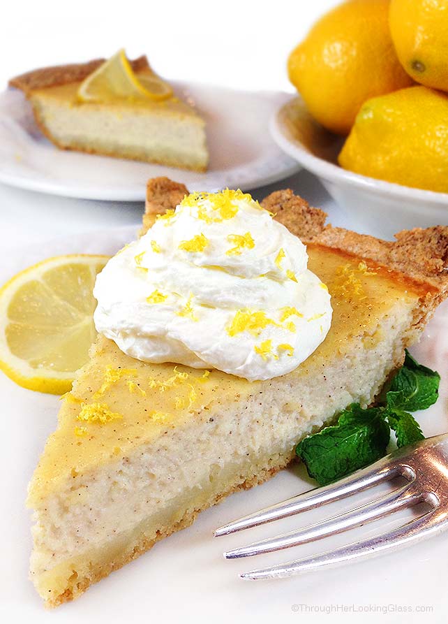 A slice of this authentic Italian ricotta pie recipe on a white plate with whipped cream, lemon zest, and fresh mint, as well as Meyer lemons and another slice of this ricotta cheesecake in the background