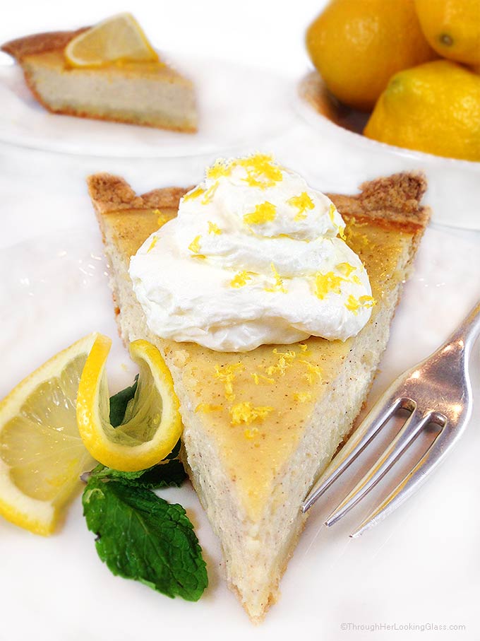 A slice of this authentic Italian ricotta pie recipe on a white plate with whipped cream, lemon zest, and fresh mint, as well as Meyer lemons and another slice of this ricotta cheesecake in the background